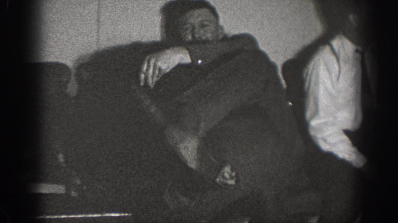 Two men relax in a domestic interior circa 1939; one reclines across an upholstered sofa while the other sits at right in a white shirt.