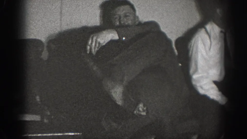 Two men relax in a domestic interior circa 1939; one reclines across an upholstered sofa while the other sits at right in a white shirt.
