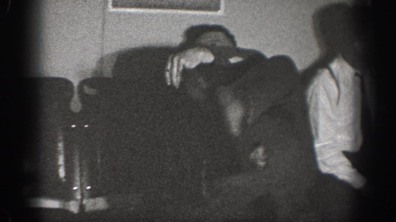 Two men in a dimly lit interior, one leaning over an upholstered sofa, black-and-white 8mm.