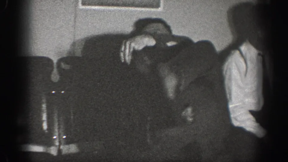 Two adult men in a dark domestic interior, ca. 1939 Chelan, Washington — one leans over an upholstered sofa while a second figure stands nearby.