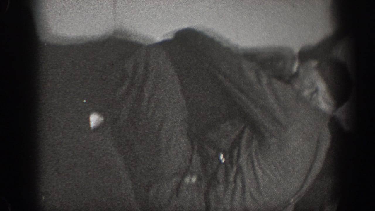 Extreme close crop on a man's torso in dark period clothing with a second figure at the frame edge; domestic interior, ca. 1939.