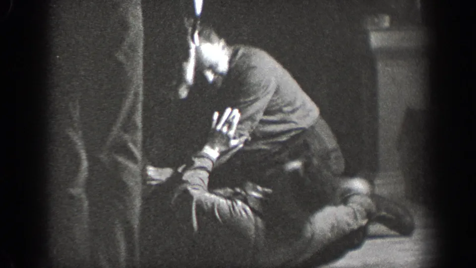 Young man crouching on an indoor wooden floor with face partially visible, background wall at right, Chelan, Washington, circa 1939.