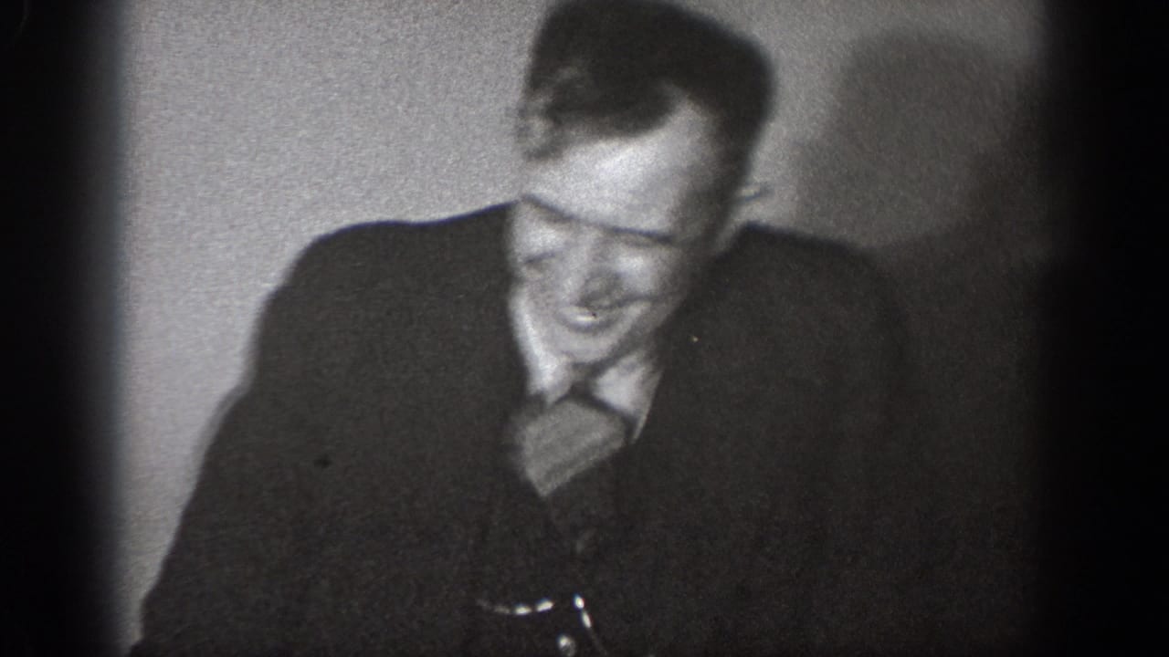 Man in dark suit and tie laughing broadly, seated against a plain light background.
