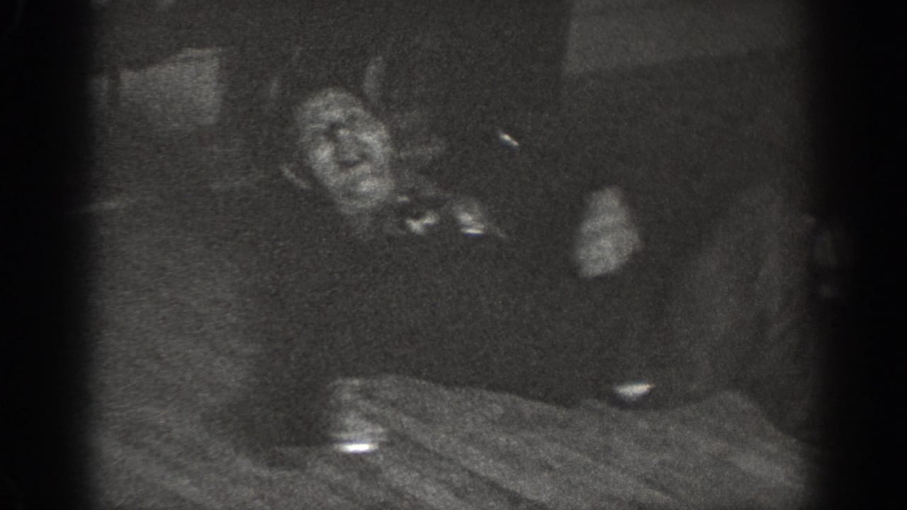 Underexposed 8mm frame showing two adults at a dining table, with motion blur and a dish or cup visible on the surface, c. 1939.