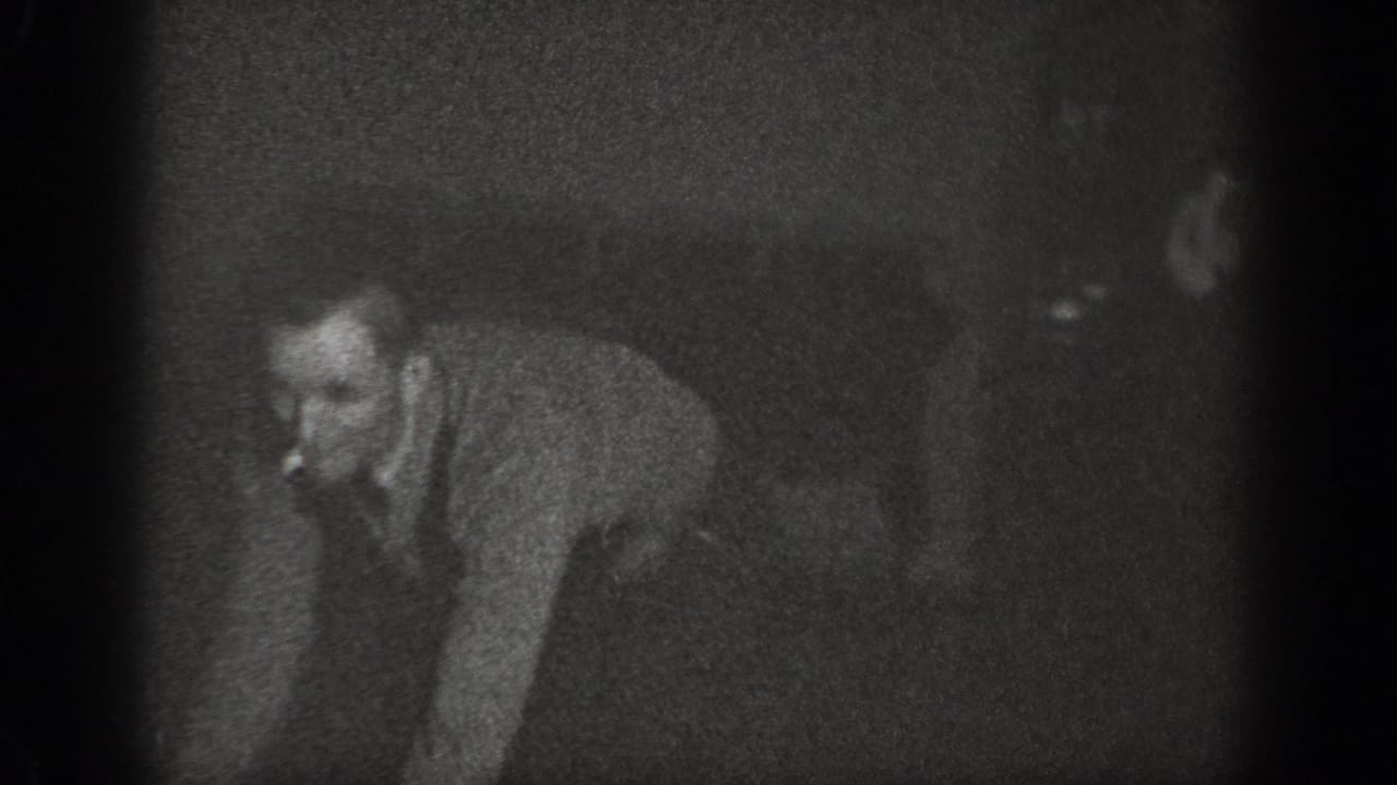 Man in collared shirt crouching on wooden floor with cigarette, second figure visible in background, dimly lit black-and-white interior.