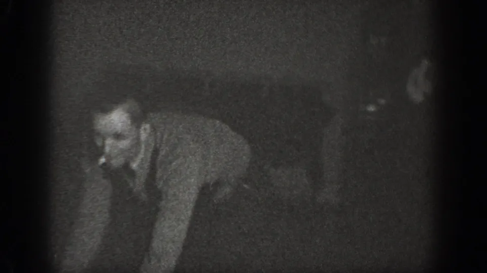 Man crouching on wood-plank floor with cigarette near mouth, another figure visible behind him; 8mm home movie footage, Chelan, Washington, circa 1939.
