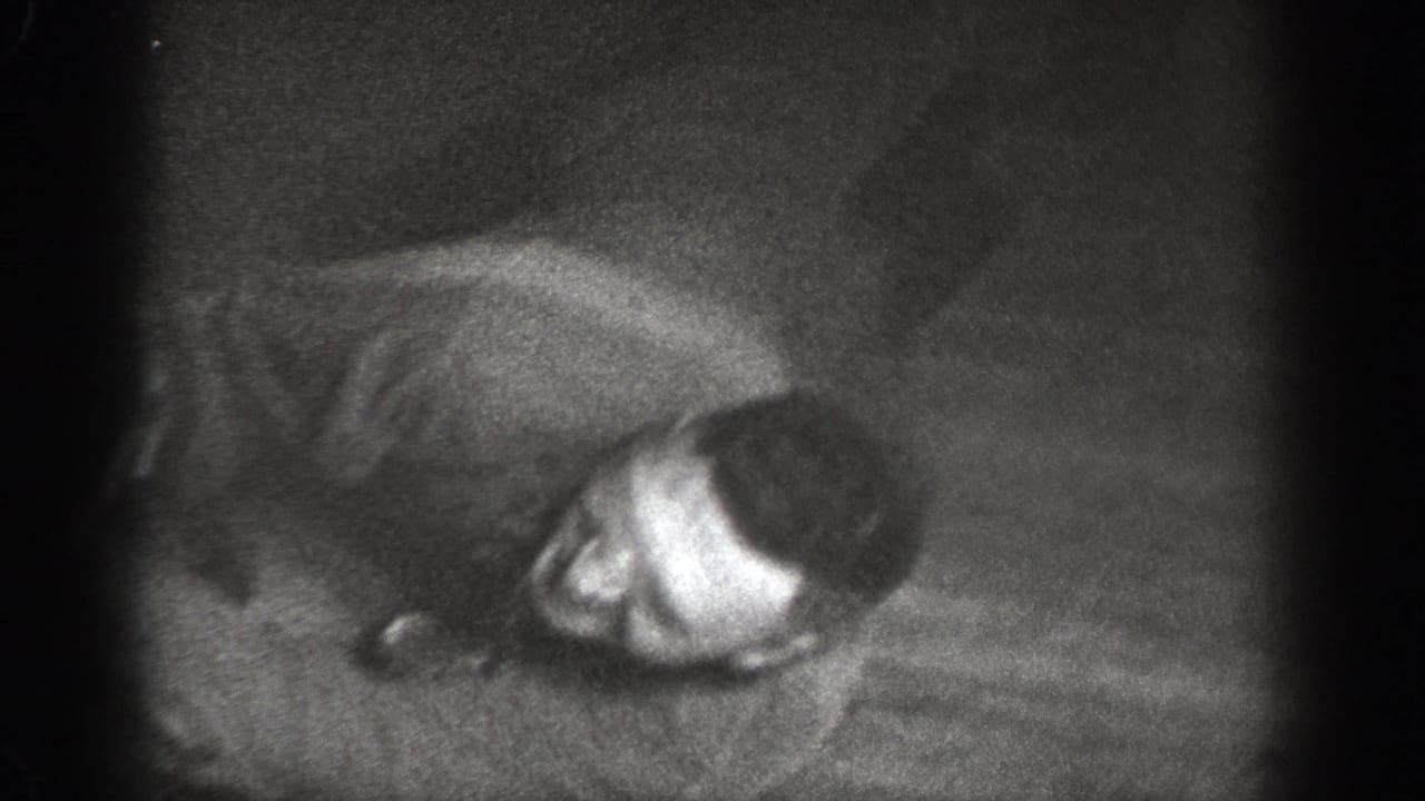 Man lying on wooden floor seen from overhead angle, relaxed or laughing, dimly lit black-and-white 8mm interior.