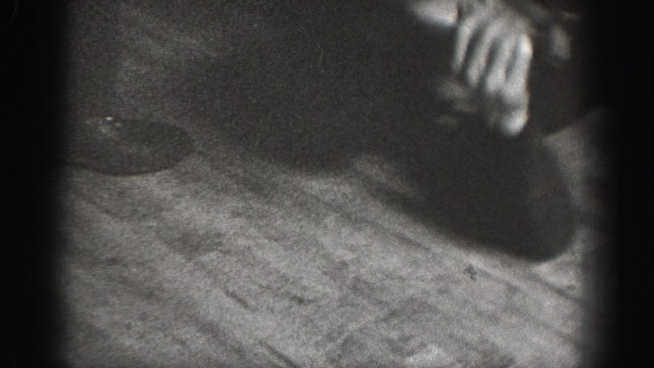 Low-angle close-up of hand reaching toward wood-plank floor; dark circular object on floor nearby; 8mm home movie footage, circa 1939.