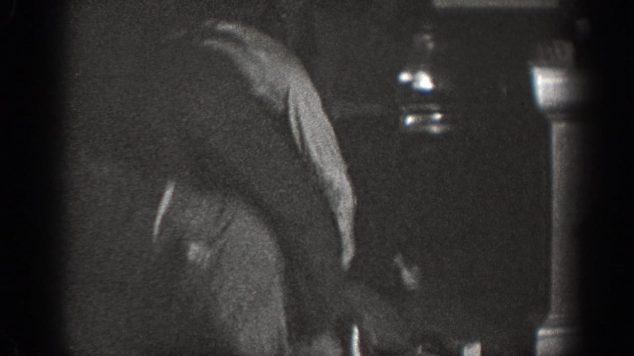 Man in dark sweater hunched forward in dim interior; two glass bottles visible on a shelf at right; 8mm home movie, Chelan, Washington, circa 1939.