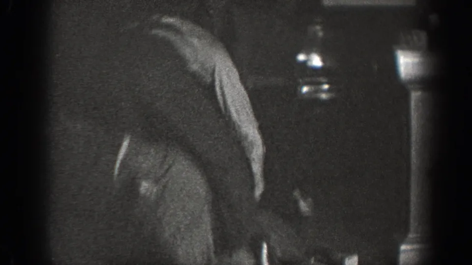 Man in dark sweater hunched forward in dim interior; two glass bottles visible on a shelf at right; 8mm home movie, Chelan, Washington, circa 1939.