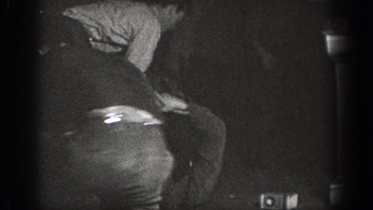 Heavily underexposed 8mm frame showing a man leaning forward in dark trousers and a light shirt, at an indoor gathering that may date to 1939 in Chelan, Washington.