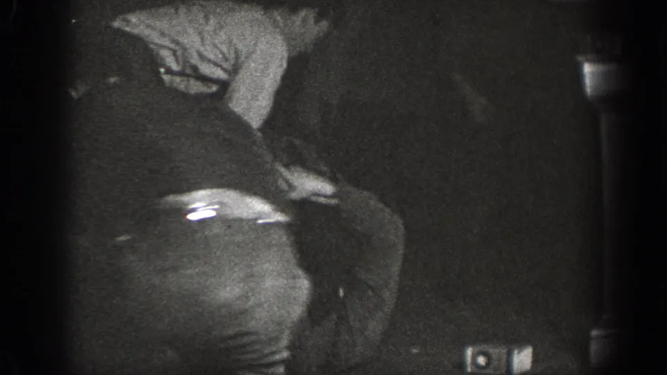 Heavily underexposed 8mm frame showing a man leaning forward in dark trousers and a light shirt, at an indoor gathering that may date to 1939 in Chelan, Washington.