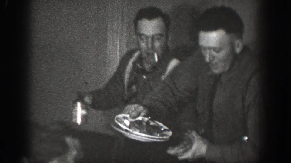 Two men smiling at an informal indoor gathering — one holding a plate of food, the other apparently smoking — in a dimly lit room consistent with a 1939 home movie from Chelan, Washington.