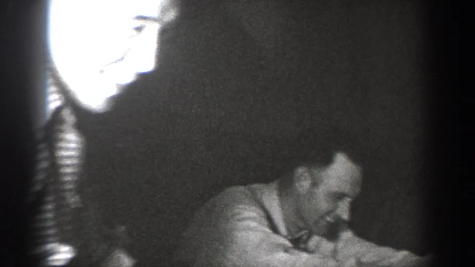 8mm frame with strong lamp overexposure at upper left illuminating a smiling man's face; a second man visible lower right at what appears to be an informal indoor gathering circa 1939.