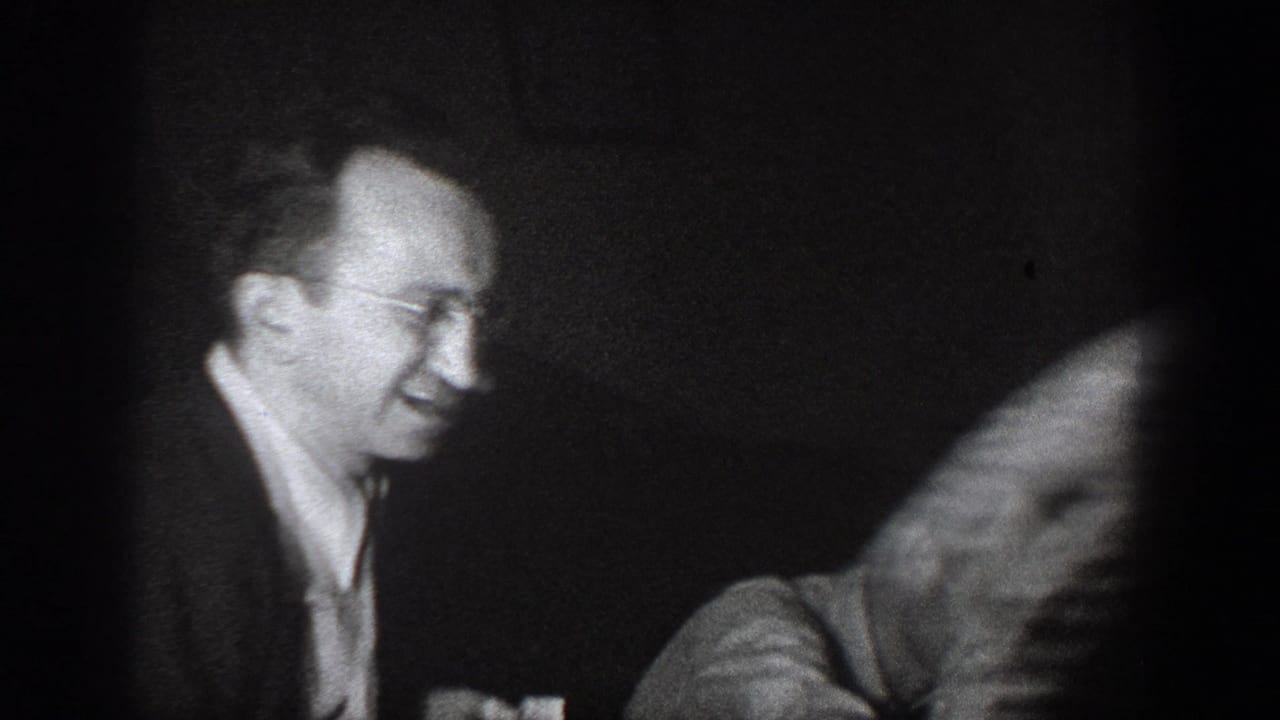 Man with wire-rimmed glasses laughing in a dark interior, second figure blurred in motion; 8mm home movie, Chelan, Washington, circa 1939.