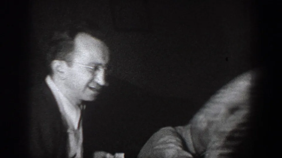 Man with wire-rimmed glasses laughing in a dark interior, second figure blurred in motion; 8mm home movie, Chelan, Washington, circa 1939.