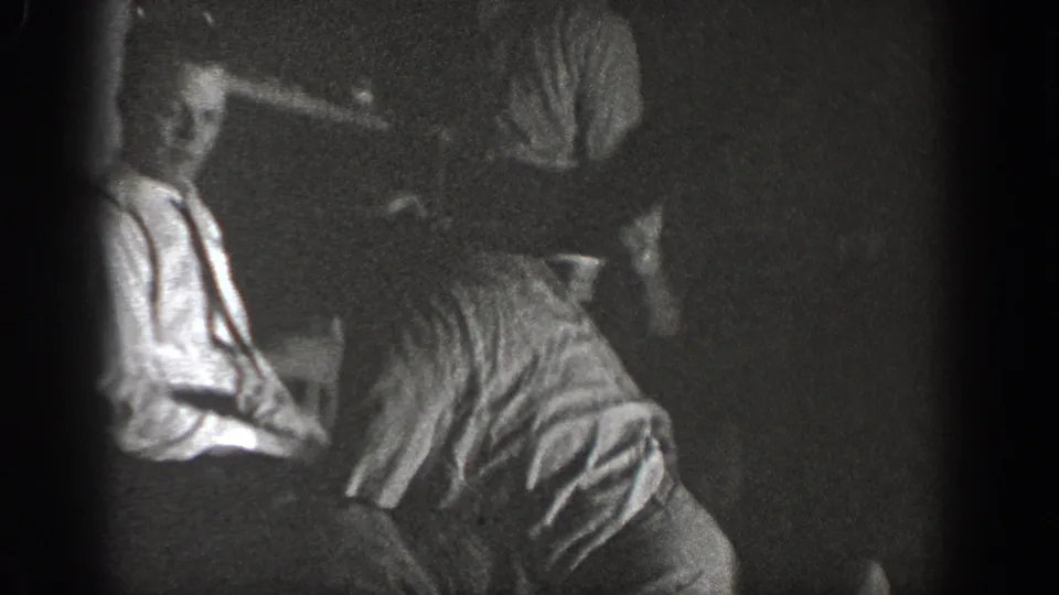 Two men in close physical contest, one facing the camera in a light shirt, recorded on 8mm in a dark interior around 1939 in Chelan, Washington.