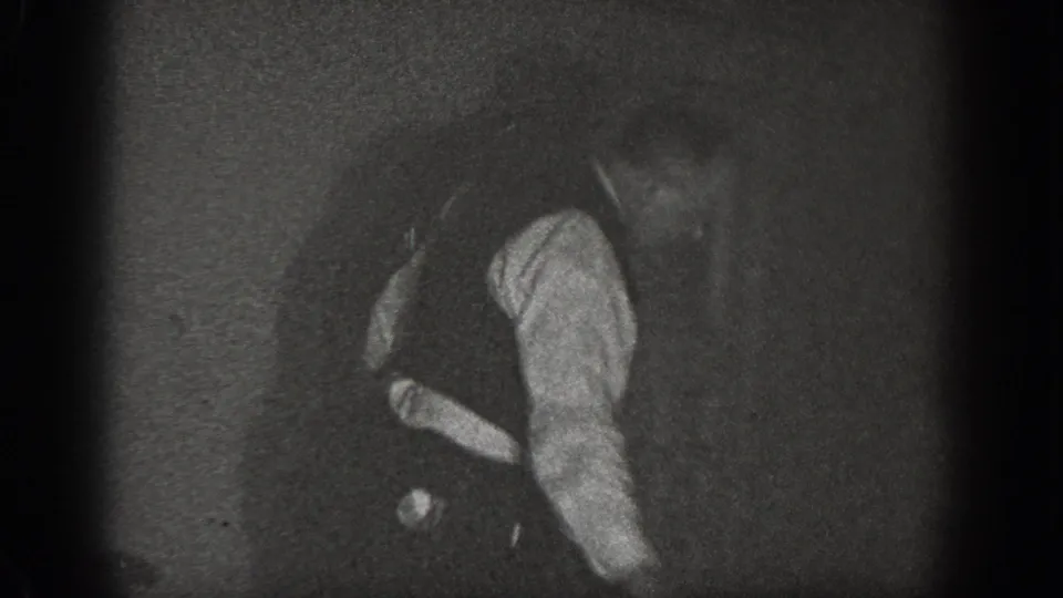 A man wearing a vest and necktie bent forward in a strenuous posture on a wooden floor, filmed on 8mm in a dark interior around 1939 in Chelan, Washington.