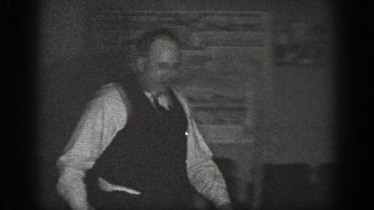 A man wearing a dark vest, white shirt, and tie stands in a domestic interior in a late 1930s home-movie frame from Chelan, Washington.