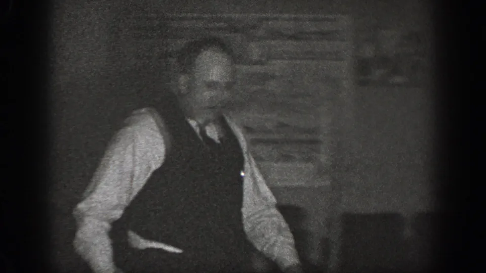 A man wearing a dark vest, white shirt, and tie stands in a domestic interior in a late 1930s home-movie frame from Chelan, Washington.