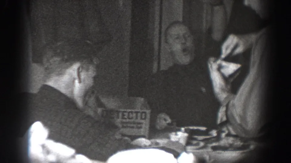Several men at a set dining table in a domestic interior, with a DETECTO-branded box visible on the table, consistent with late 1930s home-movie footage from Chelan, Washington.