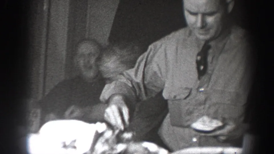 Man in a light button-up shirt and dark tie serving food at a dinner gathering in what appears to be a late 1930s domestic interior, consistent with Chelan, Washington home-movie footage.