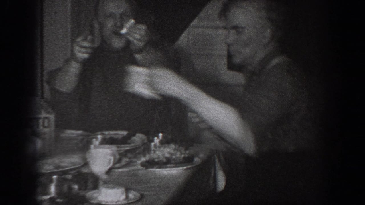 Two men at a dinner table in low light; one arm raised holding something aloft, dishes and a ceramic cup on the table.