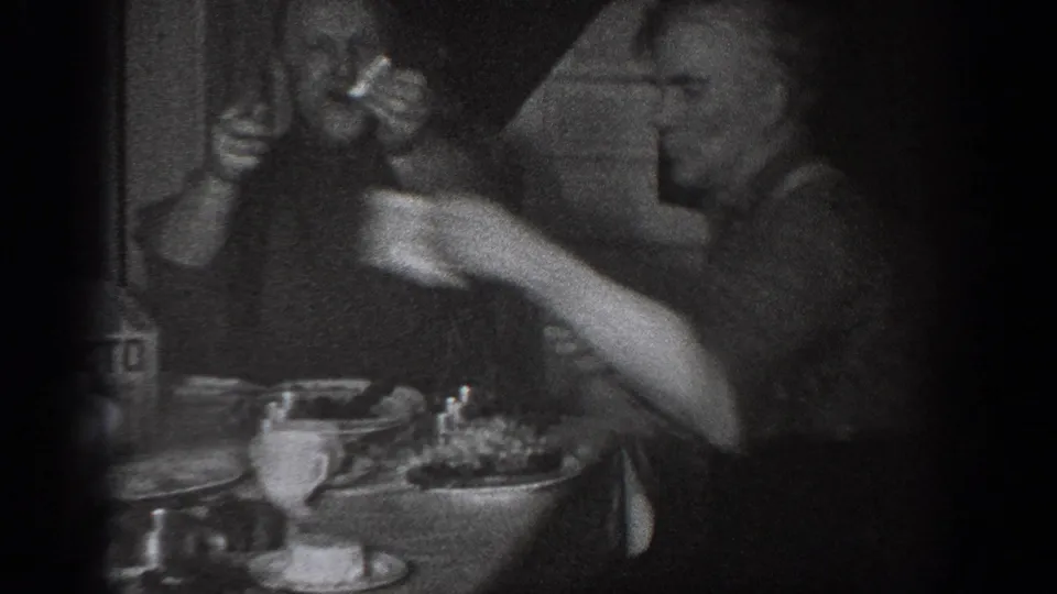 Black-and-white 8mm home movie frame circa 1939 showing two men at a set dinner table in a dimly lit interior, Chelan, Washington.
