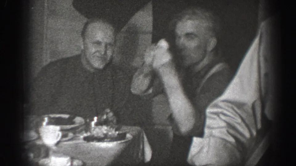 1930s 8mm home movie frame of two men at a domestic dinner table in Chelan, Washington; one man in dark clothing raises his hand.