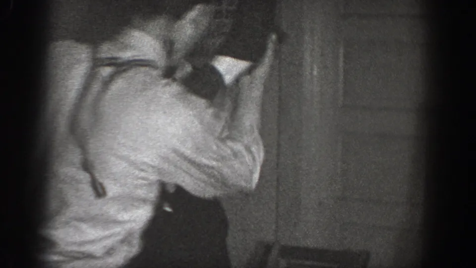 Two men roughhousing in a dim indoor setting, possibly a 1939 Chelan, Washington home; suspenders visible on one figure; 8mm film vignetting and grain throughout.