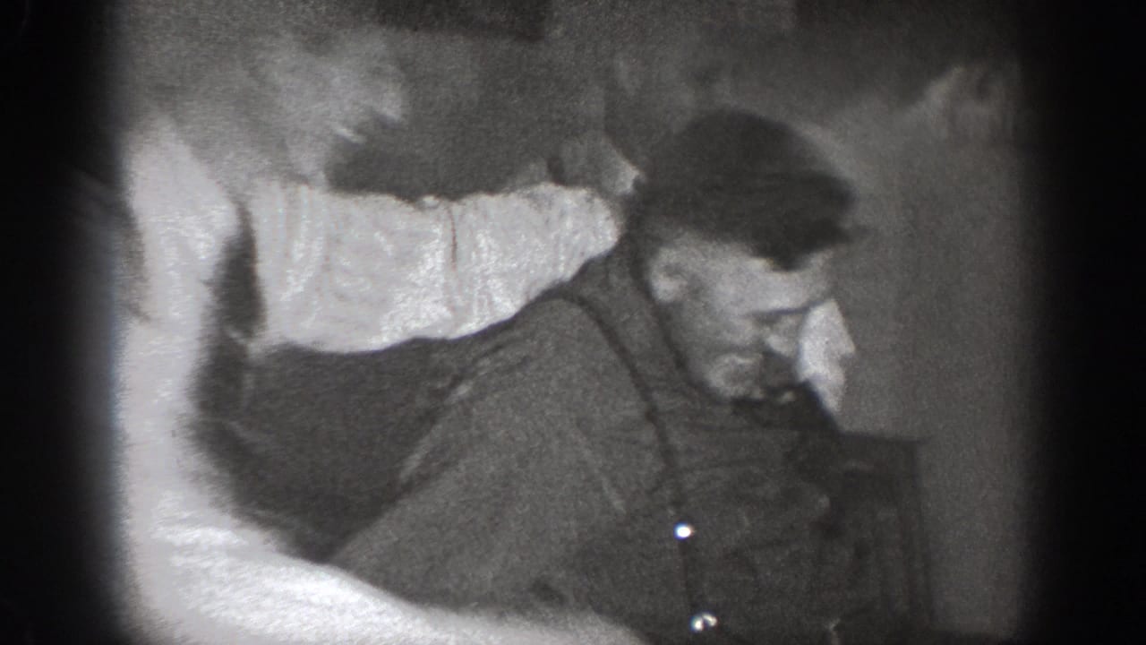 Black-and-white 8mm frame: a man in suspenders and dark work shirt leans or is pushed, with a second figure's arm visible at frame edge.