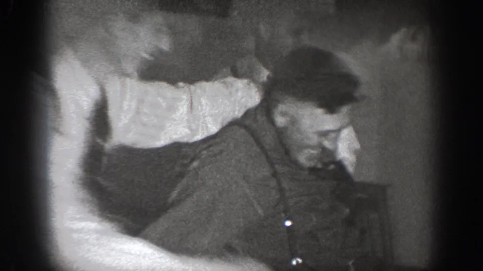 1939 8mm home movie, Chelan, Washington: man in suspenders and dark work shirt mid-struggle, second figure's arm intruding from left, dim interior.