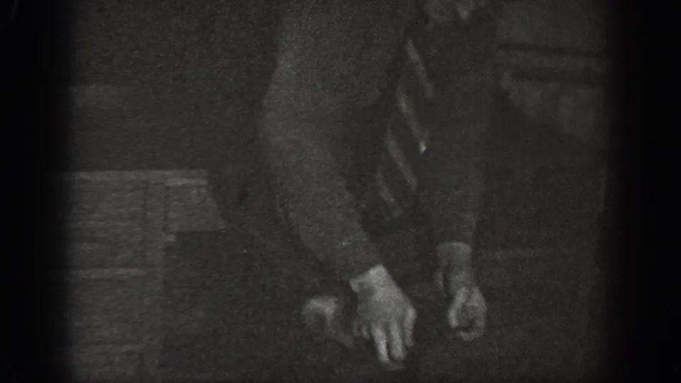 8mm home movie, ca. 1939, Chelan, Washington: figure's lower body and hands visible near wooden furniture legs on a dark interior floor.