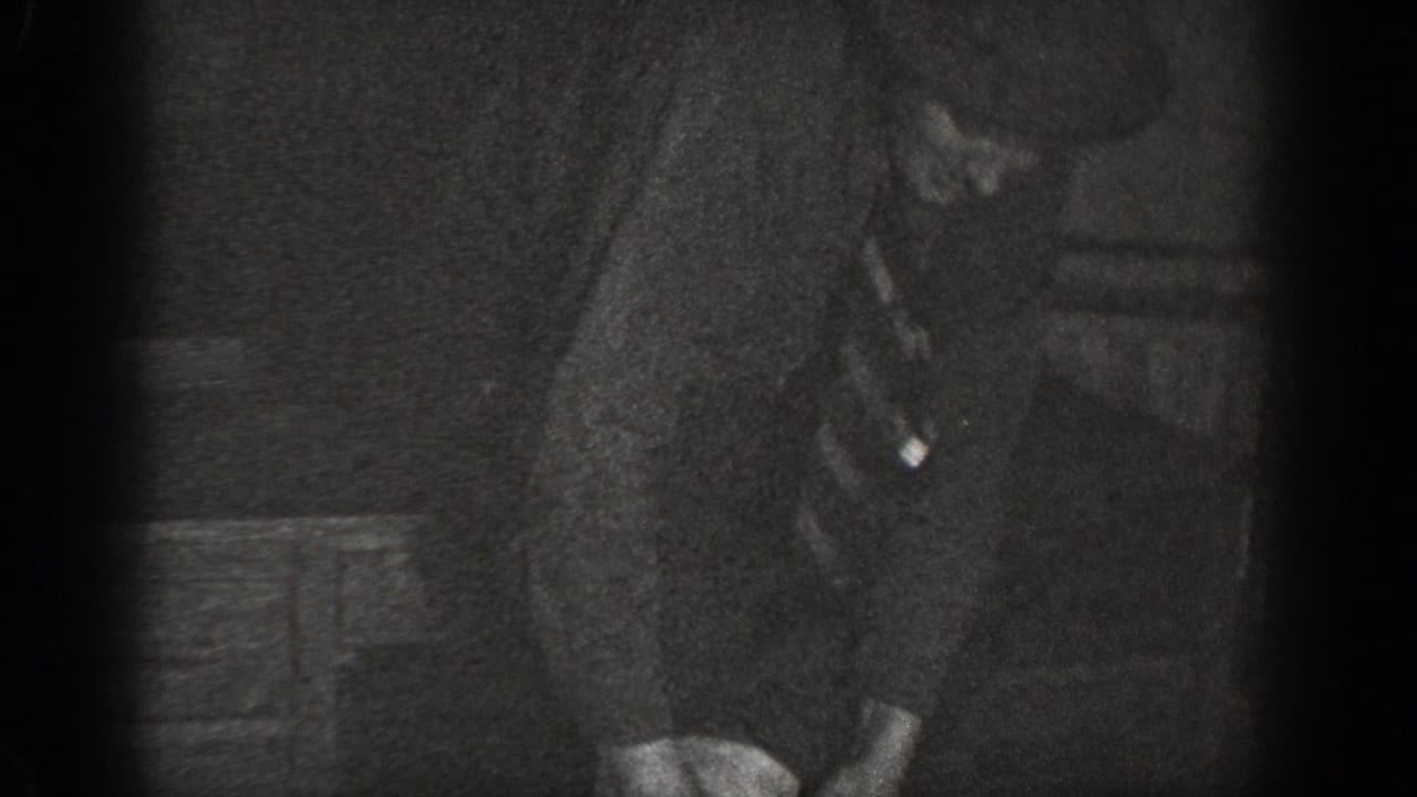 Close-up of a suited man bending at the waist in a dimly lit room, wooden crates visible in background, heavy 8mm film grain.