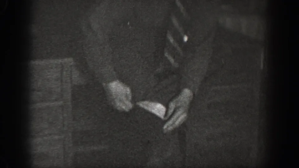8mm home movie frame showing a man in suit and striped necktie bending to handle something near foot level in a dim utilitarian interior, ca. 1939.