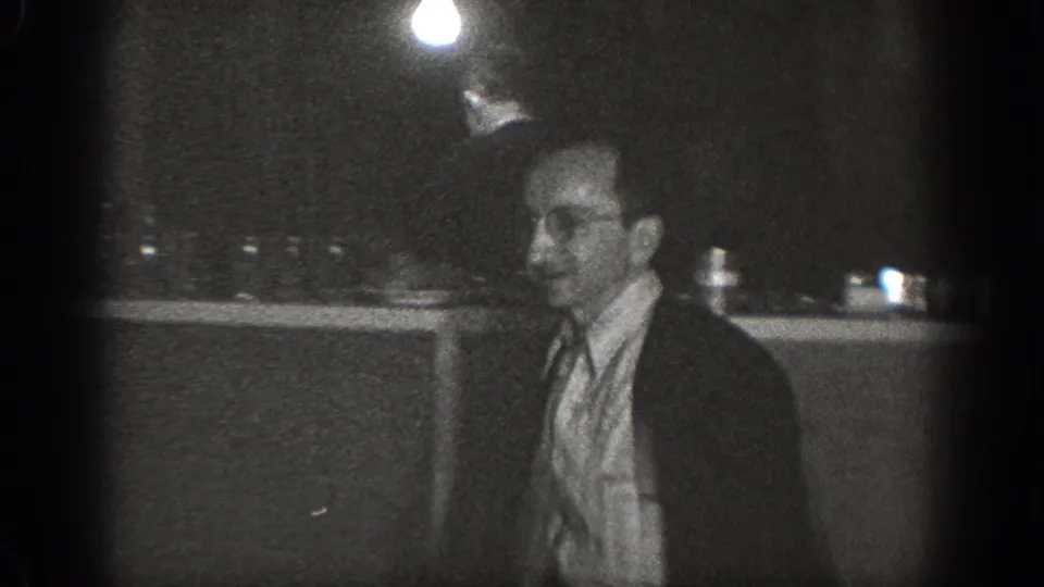 8mm home movie frame showing a suited man wearing glasses in a dimly lit back room or storage space with a bare hanging bulb and counter, Chelan, Washington, ca. 1939.