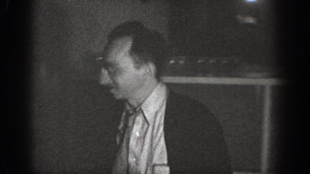 Man in collared shirt in profile view, dimly lit interior, black-and-white 8mm film.