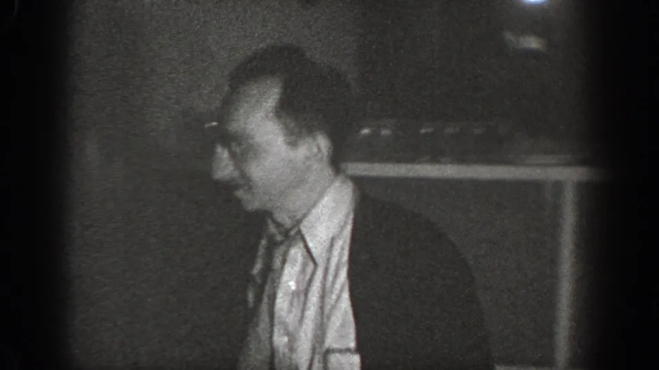 A man in a collared shirt stands in profile in a dark interior, Chelan, Washington, circa 1939, captured on 8mm home movie film.