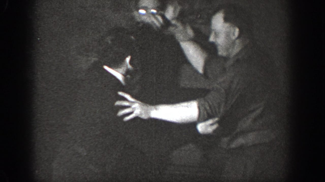 Two adult men playfully grappling in a dimly lit indoor setting, captured on 8mm home movie film, circa 1939, likely Chelan, Washington.