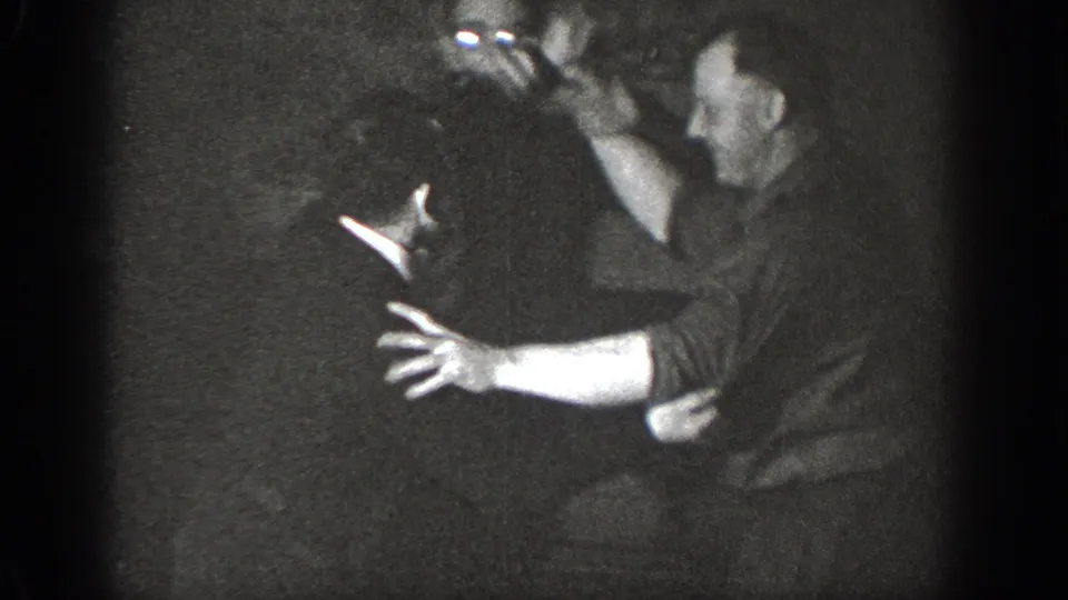 Two adult men playfully grappling in a dimly lit indoor setting, captured on 8mm home movie film, circa 1939, likely Chelan, Washington.