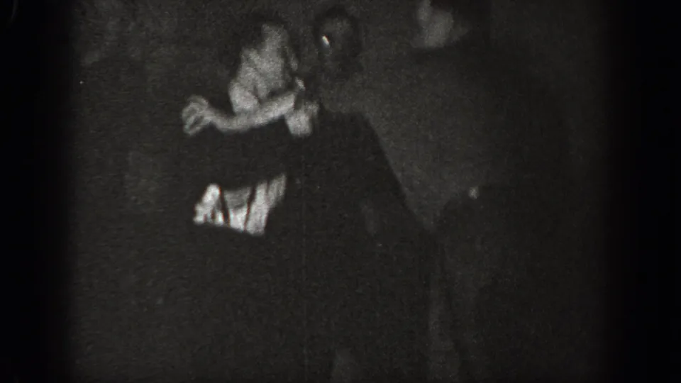 Two adult men in a playful grapple, one in a lighter-toned shirt, in a dimly lit interior from a circa-1939 8mm home movie.