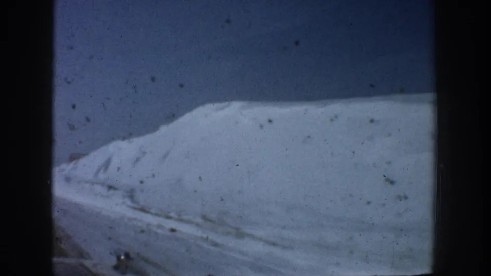 Close view of a towering roadside snowbank with textured, tracked surface against a deep blue sky, shot from a moving vehicle in Aspen, Colorado, circa 1961.
