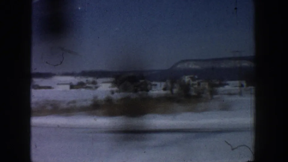 Snow-covered valley floor with a cluster of small structures in the middle distance and dark hills beyond, shot from a moving vehicle, possibly the Aspen, Colorado area circa 1961.