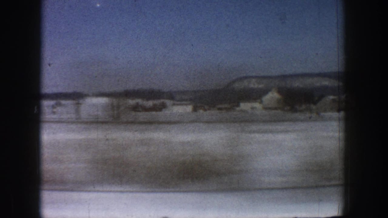 8mm moving-vehicle shot of a snow-blanketed valley with a mesa ridgeline and isolated buildings, consistent with Colorado mountain terrain circa 1961.