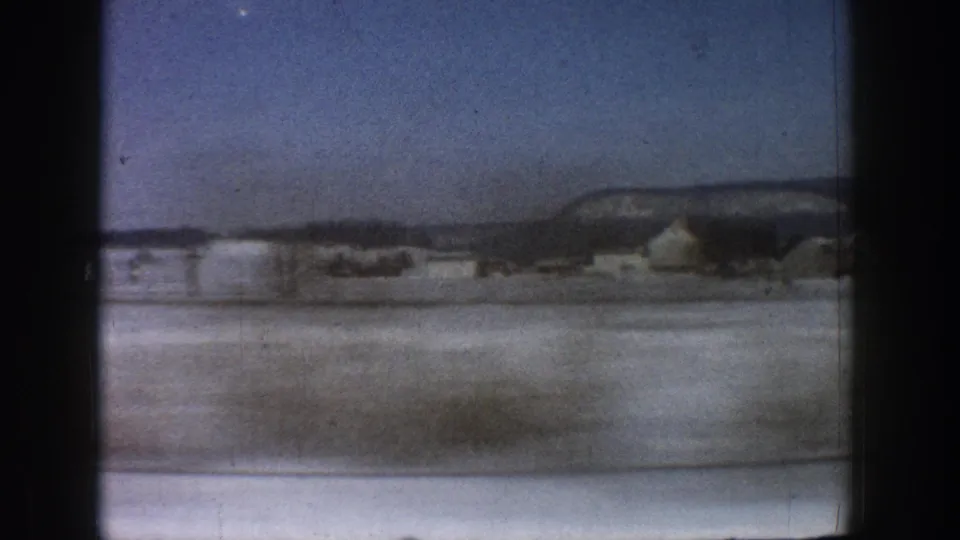 8mm moving-vehicle shot of a snow-blanketed valley with a mesa ridgeline and isolated buildings, consistent with Colorado mountain terrain circa 1961.