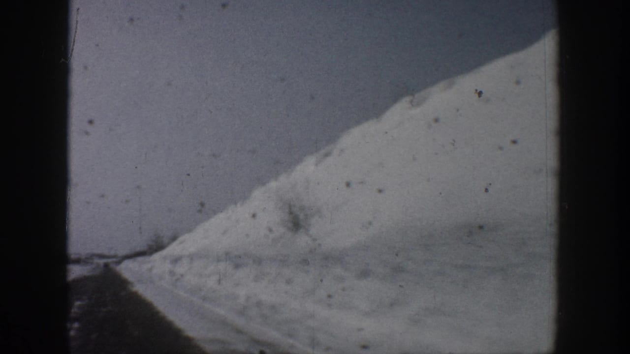 Black-and-white 8mm frame attributed to Aspen, Colorado, c. 1961, showing a steep snow-covered slope and a cleared road with deep snowbanks on either side.
