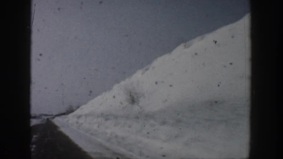 Black-and-white 8mm frame attributed to Aspen, Colorado, c. 1961, showing a steep snow-covered slope and a cleared road with deep snowbanks on either side.