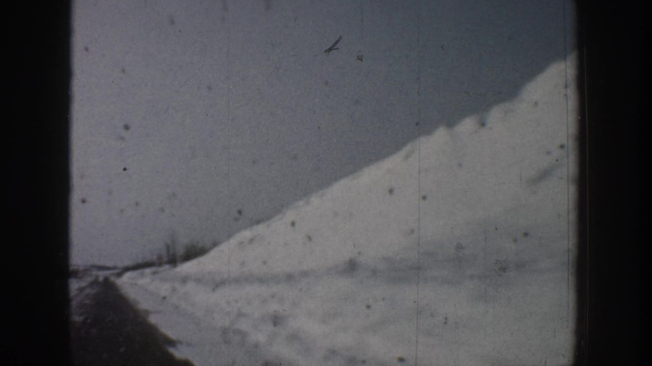 Continuation of a snow-covered hill and road sequence, early-1960s 8mm footage attributed to Aspen, Colorado, with a small airborne mark visible in the upper-right corner.