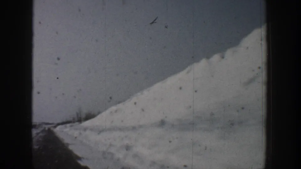 Continuation of a snow-covered hill and road sequence, early-1960s 8mm footage attributed to Aspen, Colorado, with a small airborne mark visible in the upper-right corner.