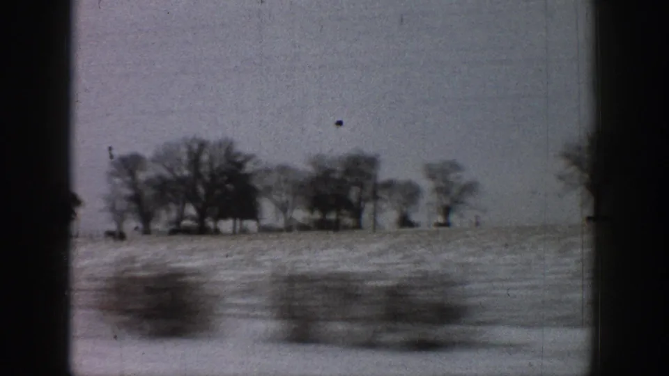 Motion-blurred foreground and bare-tree treeline across a flat winter landscape, 8mm road-trip footage attributed to Colorado in 1961, with a small airborne shape visible in the upper sky.